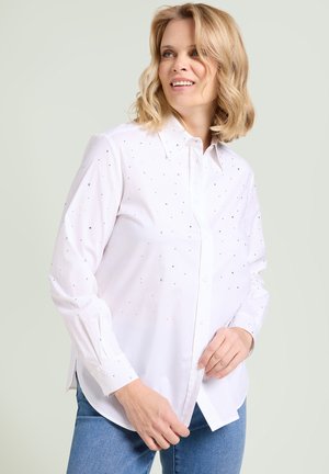 Kobieta o blond włosach do ramion, ubrana w białą koszulę zapinaną na guziki z małymi czarnymi kropkami oraz niebieskie jeansy, uśmiechająca się i patrząca w bok.