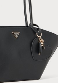 Bolsa preta texturizada com o logótipo triangular da Guess, acessórios em dourado, alças longas e um pendente em forma de coração.