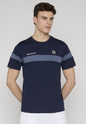 Giovane con capelli ricci corti che indossa una maglietta Sergio Tacchini blu navy con una fascia orizzontale grigia, in piedi con le mani dietro la schiena.