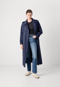 Marineblå trenchcoat med dobbeltknappede knapper, skulderepauleter og bælte. Kombinert med lyseblå jeans og hvite sneakers.