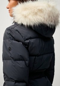 Sort pufferjakke med en foret hætte, der har et quiltet design og et broderet logo på ærmet. Blød tekstur med brede paneler.