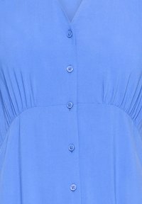 Blue fabric with five matching buttons down the center and gathered pleats on either side beneath the bust.