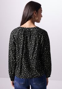 Schwarze Bluse mit weißem abstraktem Muster, langen Ärmeln, gerafftem Halsausschnitt und lockerer Passform, kombiniert mit blauen Jeans.