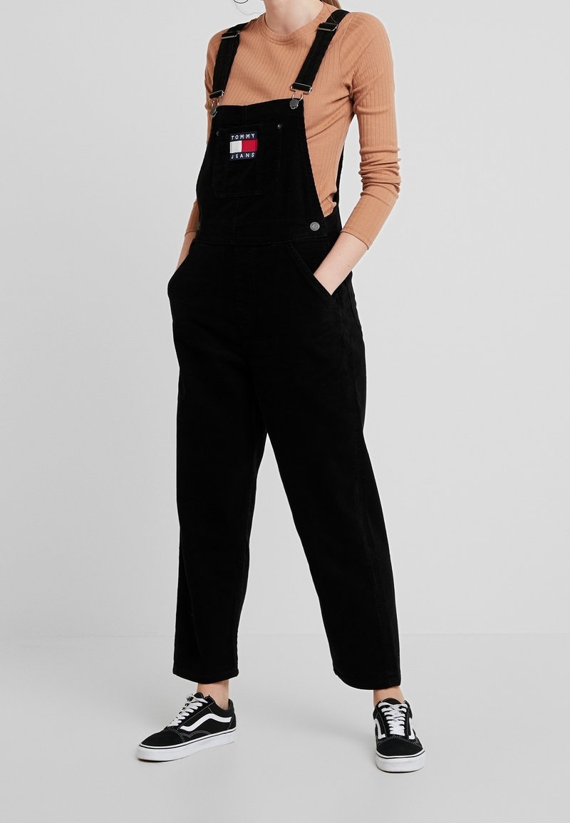 Salopette en velours côtelé noir avec des bretelles ajustables, poche avant affichant un logo coloré de Tommy Jeans, associée à des baskets noires.