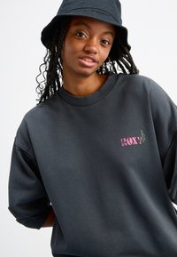 Mørkegrå hettegenser med oversized passform, ribbestrikket halskant og trykt "ROXY"-logo i rosa og grønt. Har teksturert stoff og løse ermer.