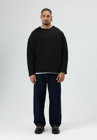 Zwarte oversized sweatshirt met verlaagde schouders, gedragen over een wit shirt met ronde hals, gecombineerd met donkerblauwe jeans met wijde pijpen en zwarte schoenen.