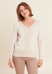 Beige V-Ausschnitt Pullover mit einem strukturierten Mittelteil, gerippten Bündchen und Saum sowie einer weichen Oberfläche. Kombiniert mit hellbraunen Hosen.