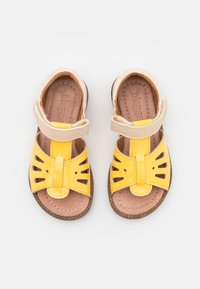 Un paio di sandali per bambini gialli e beige con cinturini in velcro, punta aperta e suole marroni cucite su sfondo bianco.