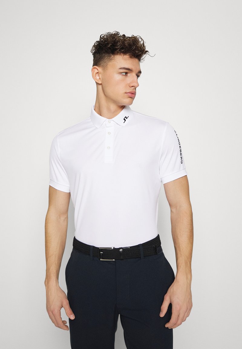 Camisa polo branca de manga curta, com três botões e um logótipo no colarinho. Usada com calças escuras e um cinto de tecido preto.