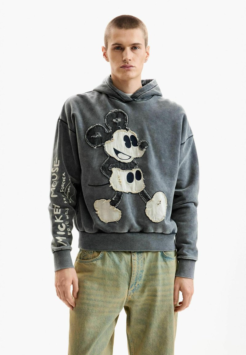 Fiatal férfi szürke kapucnis pulóverben, melyen kopott Mickey egér grafika és felirat található az ujján, világos színű farmerrel, fehér háttér előtt állva.