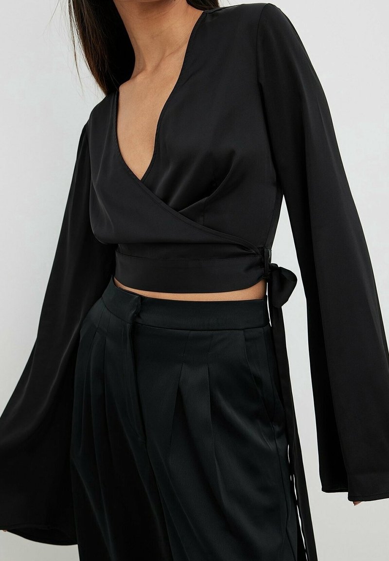 Femme portant un crop top noir en satin à manches longues et un pantalon noir plissé taille haute, sur un fond clair uni.