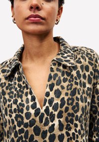 Leopardprint shirt met een kraag, voorzien van een diepe V-hals. De stof is textuur, met zwarte en bruine vlekken op een beige achtergrond.