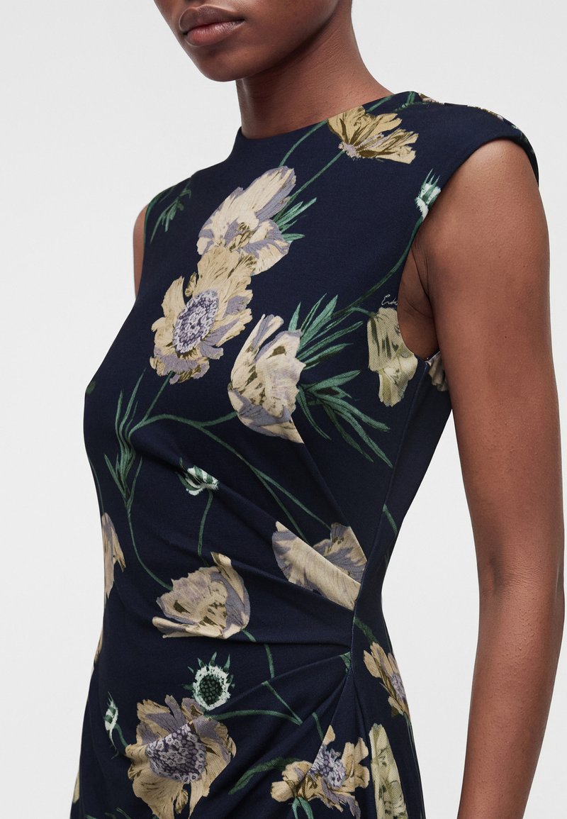 Woman wearing a sleeveless, dark navy dress with large, yellow floral print; shown from shoulder to mid-thigh against a plain background.