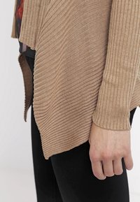 Cardigan beige à côtes avec un design fluide, présentant des manches longues et un ourlet asymétrique, fabriqué en tissu tricot doux.