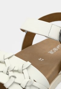 Λευκό σανδάλι με υφαντό σχέδιο λουριού και ρυθμιζόμενο πόρπη. Ομαλή υφή, ελαφριά επένδυση και καφέ πάτος με ένδειξη μεγέθους.