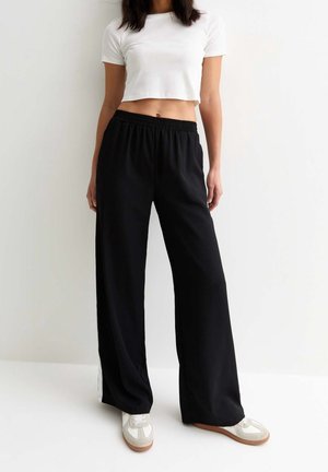 Osoba nosící bílý krátký top, volné černé kalhoty a bílé tenisky stojící před obyčejnou bílou zdí.