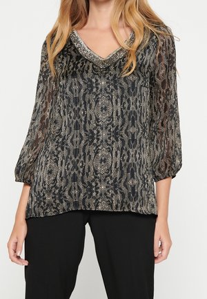 Žena oblečená v černé dlouhém rukávu s stříbrným abstraktním vzorem a ozdobným výstřihem ve tvaru V, doplněná o černé kalhoty.