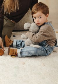 Bambino in salopette di jeans e scarpe marroni è seduto su un morbido tappeto, abbracciando un animale di peluche grigio, con uno sfondo neutro e texture.