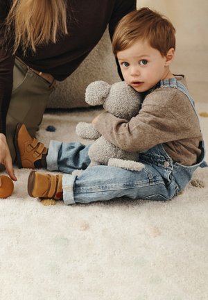 Bambino in salopette di jeans e scarpe marroni è seduto su un morbido tappeto, abbracciando un animale di peluche grigio, con uno sfondo neutro e texture.