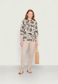 Blusa leve preta e branca com padrão paisley, calças beiges ajustadas e uma bolsa tecida em tom de castanho. Modelo está de pé sobre um fundo simples.