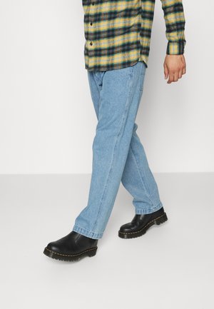 Man draagt lichtblauwe spijkerbroek, zwarte leren laarzen en een geelgroene geruite blouse met knopen, staand tegen een effen achtergrond.