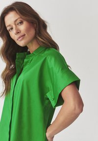 Green collared shirt, short sleeves, made of smooth fabric, featuring a button placket and stitched detailing on shoulders.