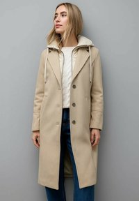Beige Wollmischungsjacke mit maßgeschneiderter Form, ausgestattet mit einer Kapuze, großen Knöpfen und Seitentaschen, getragen über einem cremfarbenen Pullover und blauen Jeans.