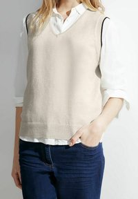 Beige Strickweste mit V-Ausschnitt, übereinander getragen über einem weißen Hemd mit Kragen. Rippbündchen und Schulterakzente in Schwarz. Kombiniert mit dunkelblauen Jeans.