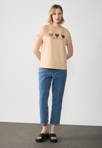 Beige kortærmet t-shirt med tre hjerte-designs, og teksten siger "BEVAR DIT VILDE HJERTE." Kombineret med blå jeans og sorte slip-on sko.