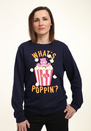 Mørkeblå sweatshirt med et popcorn grafik med en bjørn, multikolorede tekst "HVAD SKER DER?", og hvide popcorn detaljer.