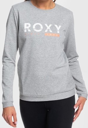 Femme vêtue d'un sweat-shirt gris à manches longues avec le texte "ROXY 1990" en blanc et orange, associé à un pantalon noir.