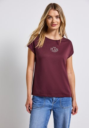 T-shirt corto marrone in cotone, con scollo rotondo, vestibilità ampia e logo in testo bianco sulla parte anteriore. Indossato con jeans blu.