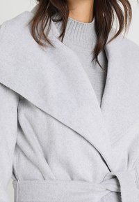 Ljusgrå omslagskappa gjord av mjukt, strukturerat tyg, med bred krage, långa ärmar och ett matchande bälte i midjan.