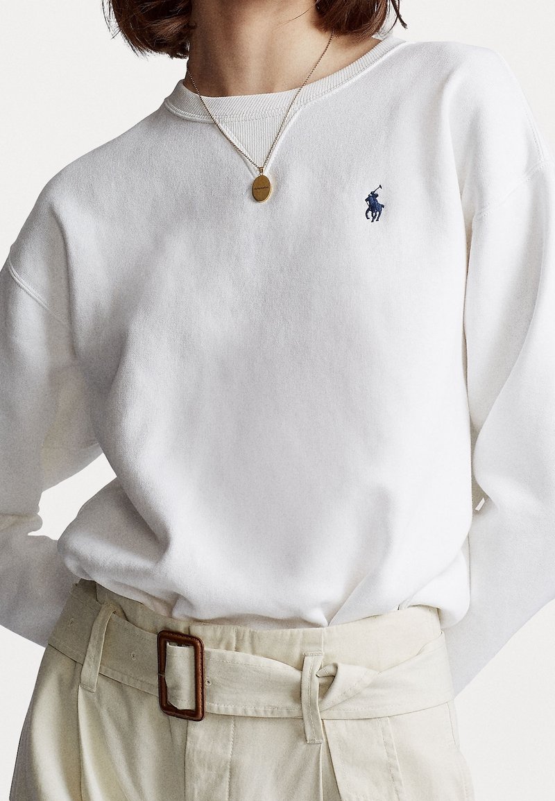 Kvinna som bär en vit långärmad skjorta med en marinblå polo-spelare-logotyp, beigefärgade högmidjade byxor med rektangulär spänne, och en guldpendelhalsband.