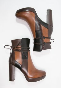 Een paar vrouwen enkellaarsjes met hoge hakken in bruin en zwart leer, met een zijrits en vetersluiting op een witte achtergrond.