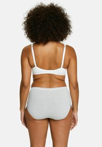 Donna con capelli ricci che indossa un reggiseno bianco e mutandine grigie a vita alta, vista di spalle contro uno sfondo neutro.