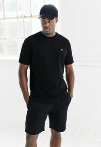 Must must t-s�ärk lühikeste varrukatega ja väikese logoga vasakul rinnal, koos mustade lühikesed pükstega, kandmisega musta mütsiga.