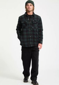 Chemise en flanelle à carreaux noirs et verts avec deux poches poitrine, boutons, et une coupe décontractée, associée à un pantalon noir et un bonnet tricoté vert.