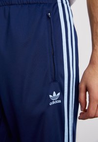Jūras spēku sporta bikses ar gaiši zilu trīs svītru dizainu, elastīgu jostu un rāvējslēdzēju kabatu ar izšūtu logotipu augšstilbā.