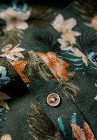 Tessuto floreale verde con fiori arancioni e blu, dotato di chiusura a bottone e superficie testurizzata e tessuta.