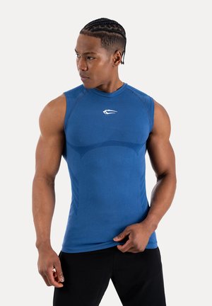 Uomo muscoloso con capelli intrecciati che indossa una maglietta atletica blu aderente senza maniche e pantaloni neri, che guarda verso sinistra.