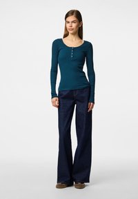 Turkos ribbad långärmad tröja med rund halsringning och knapppdetaljer, kombinerad med mörkblå vida jeans och bruna skor.