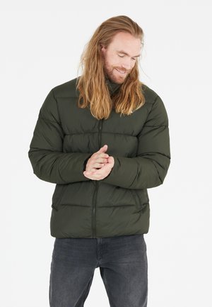 Grøn oppustelig jakke med lynlåse foran, syede sektioner og høj krave. Parret med mørke jeans ser stoffet glat og isoleret ud.