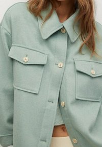Giacca oversized di colore verde chiaro, in tessuto morbido, con due tasche sul petto, caratterizzata da bottoni beige e un design colletto classico.
