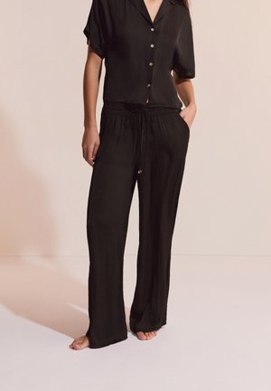 Frau trägt ein locker sitzendes, schwarzes, kurzärmliges Hemd mit Knöpfen und passende, weit geschnittene Hose mit Kordelzug, steht barfuß vor einfachem Hintergrund.