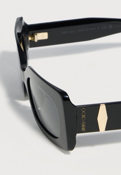 Μαύρα γυαλιά ηλίου με τετράγωνο πλαίσιο και σκουρόχρωμους φακούς, με χρυσές γεωμετρικές λεπτομέρειες στο πλάι, με τη σήμανση "Jimmy Choo" σε χρυσά γράμματα.