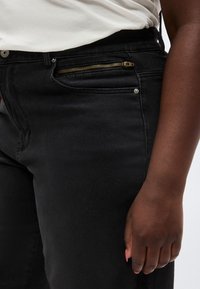 Κοντινό πλάνο ενός ατόμου που φοράει μαύρα τζιν με χρυσό φερμουάρ στην μπροστινή τσέπη και λευκό πουκάμισο φυλαγμένο μέσα.