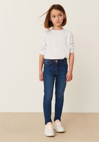 Plave traperice u kombinaciji s bijelom majicom dugih rukava, s okruglim izrezom i podvrnutim rukavima. Outfit je upotpunjen bijelim tenisicama.