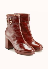 Bottines en cuir marron avec une finition brillante, à bout carré et talon épais. Présente une sangle enroulante avec une boucle dorée circulaire.