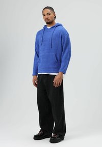 Blauwe zachte hoodie met een voorzak en trekkoord; gedragen over een wit shirt en gecombineerd met zwarte wijde broekspijpen en zwarte schoenen.